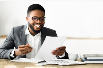 Poster - A man in a suit is sitting at a desk, reviewing documents. He is smiling and holding a pen in his hand.
