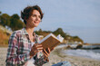 © ViDi Studio - Side profile view young fun happy dreamful woman wear shirt casual clothes reading paper book novel sit rest on sea ocean sand shore beach outdoor seaside in summer day free time. Lifestyle concept.