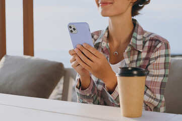 Wall Mural - Close up cropped young happy woman wear shirt casual clothes sitting alone at street cafe table in coffee shop drink tea hold in hand use mobile cell phone rest in restaurant during free time outdoor.