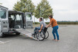 © 24K-Production - Female assistant helping a male person in wheelchair with transport using accessible vehicle van ramp. Disability and mobility concepts.