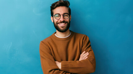 Wall Mural - Young handsome man with beard, wearing a casual sweater and glasses, smiling with crossed arms, looking confidently at the camera against a blue background