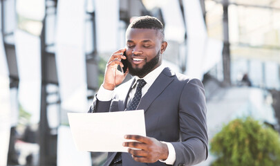 Wall Mural - Phone Negotiations. African Entrepreneur Talking On Cellphone Holding Documents And Papers Standing In City Urban Area. Copy Space, Panorama