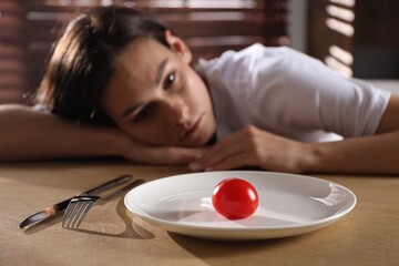 Canvas Print - Eating disorder. Sad woman at wooden table with cutlery, tomato and plate indoors