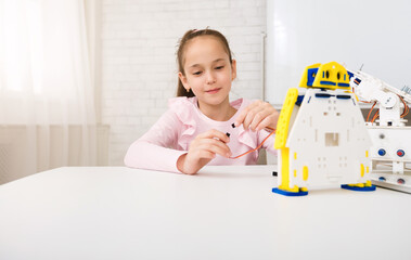 Poster - A young girl sits at a white table, focused on assembling a robot kit. She holds a small wire in her hand, carefully connecting it to the robots wiring. The robot is made of white plastic