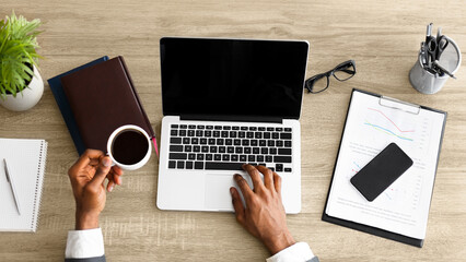 Poster - Office workplace top view. Black businessman working at wooden desk, using laptop and drinking coffee