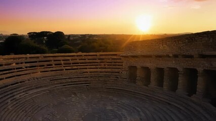 Sticker - A dramatic view of an ancient amphitheater now in ruins with the sun setting in the background.