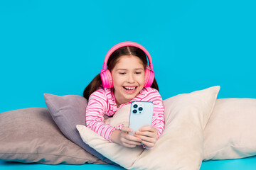 Poster - Photo of funny girl dressed striped shirt in headphones lay on pillows play video game on smartphone isolated on blue color background