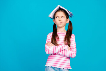 Poster - Photo of minded small girl dressed striped shirt holding book on head arms crossed look empty space isolated on blue color background