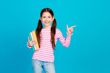 Poster - Photo of clever small girl with ponytails dressed striped shirt holding book directing empty space isolated on blue color background