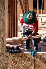 Wall Mural - Construction worker in toolbelt and sun hat working with a pneumatic nail gun on new house wood construction framing, summer construction season
