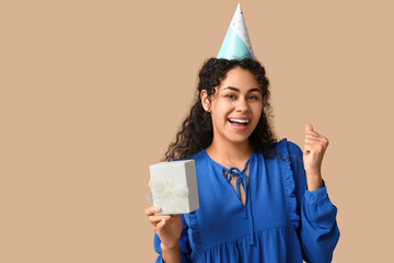 Poster - Beautiful young African-American woman in party hat with gift box on brown background