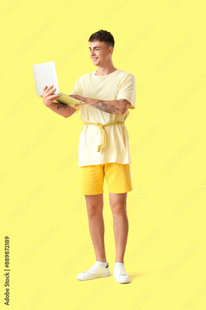 Young man with tape measure using laptop on yellow background. Weight loss concept