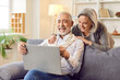 © Studio Romantic - Elderly couple is spending leisure time together, using a laptop and laughing, sitting on a sofa in the living room at home. They are engaged in online entertainment, enjoying internet activities.