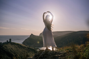 Wall Mural - A woman stands on a hill overlooking the ocean. She is wearing a white dress and has her hands on her head. The scene is serene and peaceful, with the woman looking out over the water.