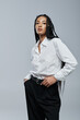 © LIGHTFIELD STUDIOS - A young woman in a white shirt and black pants poses against a grey backdrop. She has black braided hair and a confident expression.