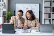 © Wasana - A man and a woman are sitting at a desk with a laptop and a tablet