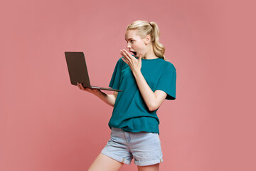 Young attractive woman dressed casual attire looking surprisingly, with shock to laptop screen against pastel pink studio background. Concept of online communication, technology, shopping, sale .