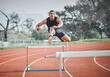 © DavisShared/peopleimages.com - Hurdles, fitness and man sprinting on track at stadium training for race, competition or marathon. Energy, jump and male athlete running by equipment for cardio workout or exercise at sports arena.