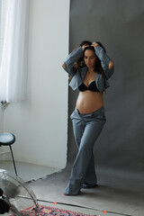 A beautiful pregnant brunette woman in a cropped gray stylish suit on a gray paper background in the studio. A pretty girl with a bare stomach in a fashionable suit is sitting barefoot on a gray backg