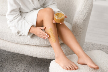 Poster - Young woman in bathrobe applying body scrub on legs on white sofa at home, closeup