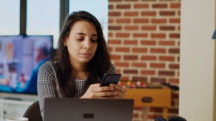 Smiling person checking mobile phone notifications at computer desk, taking break from work. Indian woman in cozy apartment pausing job tasks to read messages on cellphone, camera B close up