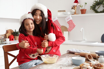 Wall Mural - Happy Asian mother hugging her little daughter while cooking cinnamon rolls in kitchen on Christmas eve