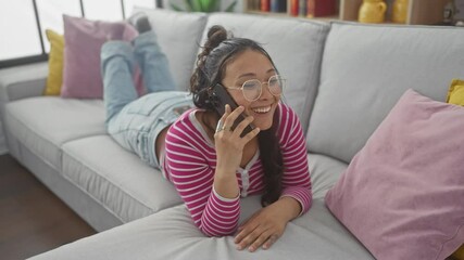 Poster - A cheerful young hispanic woman talking on a smartphone while relaxing on a couch in a cozy living room.