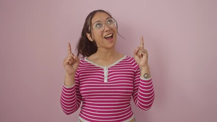 Wall Mural - Surprise, joy on the face of a young, hispanic woman in a striped t-shirt. she's pointing upward, looking amazed. all over a pink, isolated background.