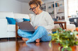 © simona - Cheerful woman using laptop on her legs sitting crossed legs on the floor at home in living room. Business and online job. Searching for on the net. Female smiling and writing on notebook alone