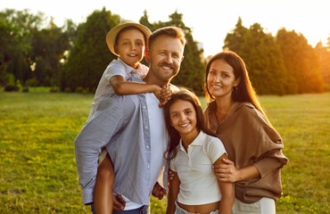 Wall Mural - Portrait of happy smiling family of four with son and daughter standing in the summer park and looking cheerful at camera. Parents with two kids walking in nature enjoying sunny day together.