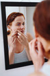 © peopleimages.com - Woman, skincare and mirror for pimple in home, face inspection and acne problem for wellness in bathroom. Breakout, pinch and upset for beauty or dermatology, blackheads and checking sebum production