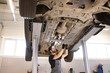 © sofiko14 - Mechanic performing maintenance and repair work under raised car in auto repair shop. Worker wearing gloves and focusing on vehicle service. Professional garage environment with car on hydraulic lift.