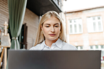 young beautiful blond caucasian woman sitting at table in outdoor terrace of cafe in summer day with notebook, businesswoman working, freelancer typing text and smiling