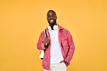 Happy young adult African man hipster standing isolated on yellow background looking at camera. Smiling cool generation z Black student guy wearing backpack headphones posing for portrait.