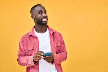 Young happy African Black man user with smartphone in hands standing isolated on yellow background holding cellphone using mobile cell phone looking at copy space advertising betting app.