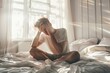 © Igor - A young man sitting in a bed with a headache holding head in hands, morning light pours from the windows