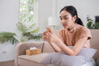 © wichayada - Young Woman Sitting on Sofa Holding Birth Control Pills in a Modern Living Room Setting