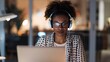 © Nuchylee - Woman wearing headphones works late on her laptop in a modern office, dedication and concentration in a professional setting.