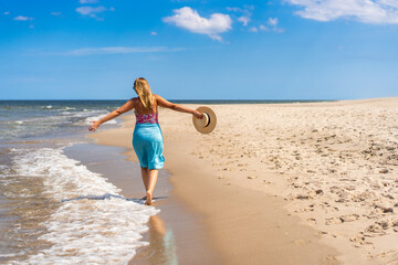 Wall Mural - Summer vacation. Beautiful young woman in colorful swimsuit and blue pareo walking with hat in her hand on the sandy beach on a beautiful sunny day. Back view