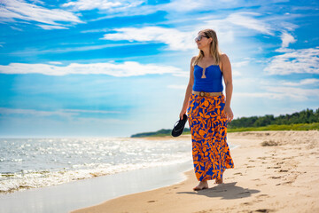 Wall Mural - Summer holidays on beach by Baltic Sea . Beautiful young woman in blue swimsuit and patterned skirt standing on white sandy beach holding dark blue flip-flops in her hand on sunny day. Front view