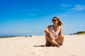 Wall Mural - Summer holidays. Portrait of young beautiful woman with long blonde hair in light dress and sunglasses sitting on ground on sandy beach on sunny day. Energy, joy of life and youth