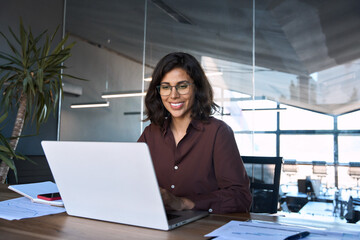 Smiling 30s latin hispanic middle-aged business woman working on laptop computer in modern office. Indian young businesswoman professional employee using pc doing online banking analysing at workplace