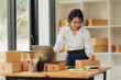 © Kritdanai - Successful Asian businesswoman selling packages online, smiling, working alone at table with laptop.
