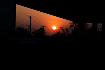 Sunset with a large orange sun partially visible on the horizon, silhouetted trees and power lines in the foreground, darkened landscape with a slight overhang in the upper part of the image