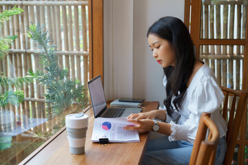 Young asian businesswoman checking marketing data of business on document and typing data on keyboard laptop while thinking about investment and planning of new startup project for working in cafe