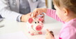 © H_Ko - Hand of little girl putting coins in funny piggy bank pin slot closeup