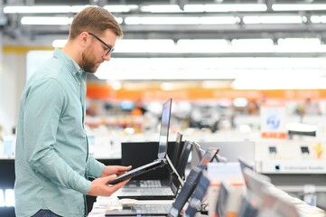 Wall Mural - A young man stands behind his laptop at the electronics store. A young man chooses a laptop in the store