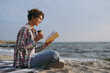 © ViDi Studio - Full body side view young woman she wear shirt casual clothes reading paper book novel drink coffee sits rest on sea ocean sand shore beach outdoor seaside in summer day free time. Lifestyle concept.