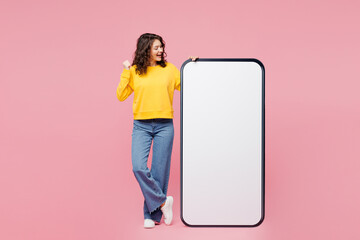 Wall Mural - Full body young woman wear yellow sweatshirt casual clothes big huge blank screen mobile cell phone smartphone with area do winner gesture isolated on plain pastel pink background. Lifestyle concept.