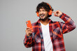 © ViDi Studio - Young happy excited Indian man he wearing red shirt casual clothes listen to music in headphones use hold mobile cell phone isolated on plain grey color background studio portrait. Lifestyle concept.